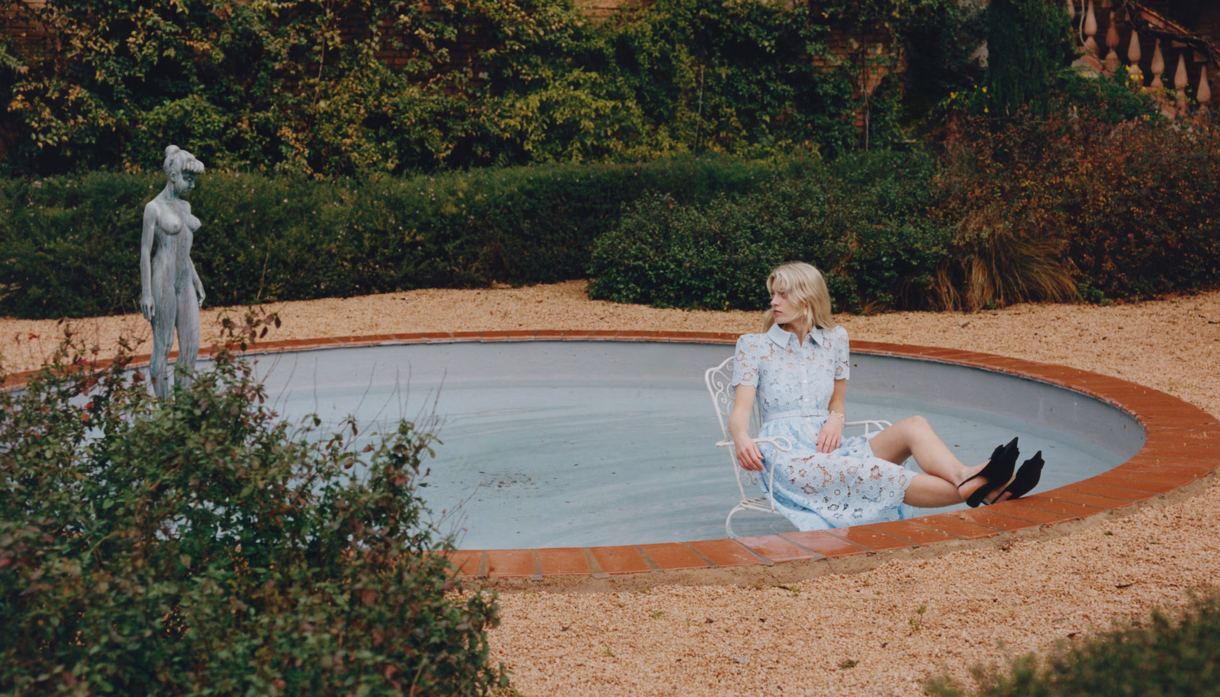 Woman in a blue lace dress sitting by a pool with a statue in the background.