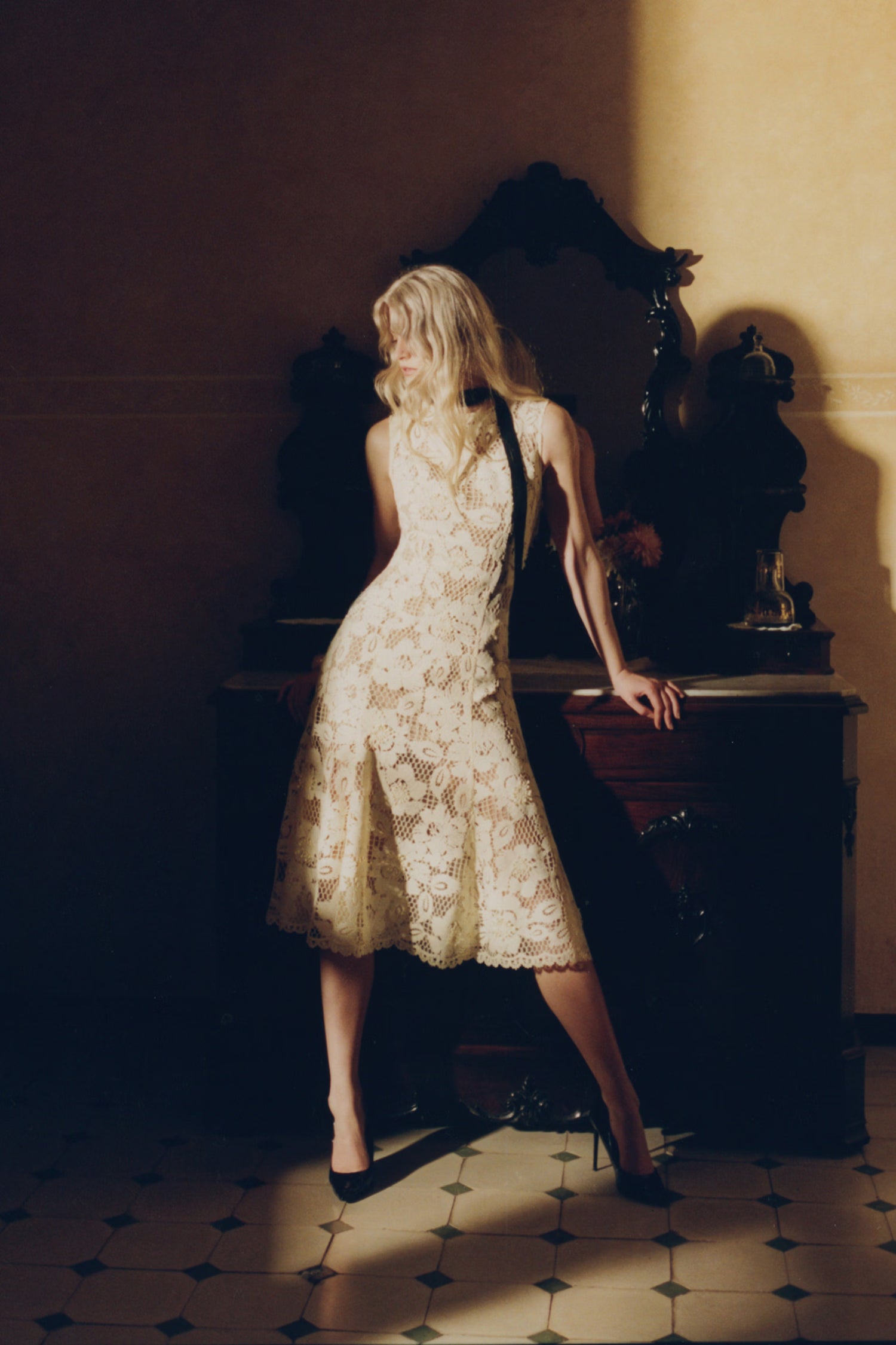 Woman in a yellow lace dress standing in a dimly lit room with a wooden dresser.