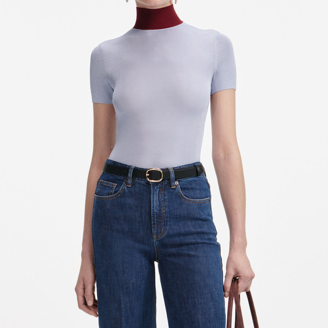 Woman in light blue short-sleeve top with red turtleneck, dark blue jeans, black belt with gold buckle