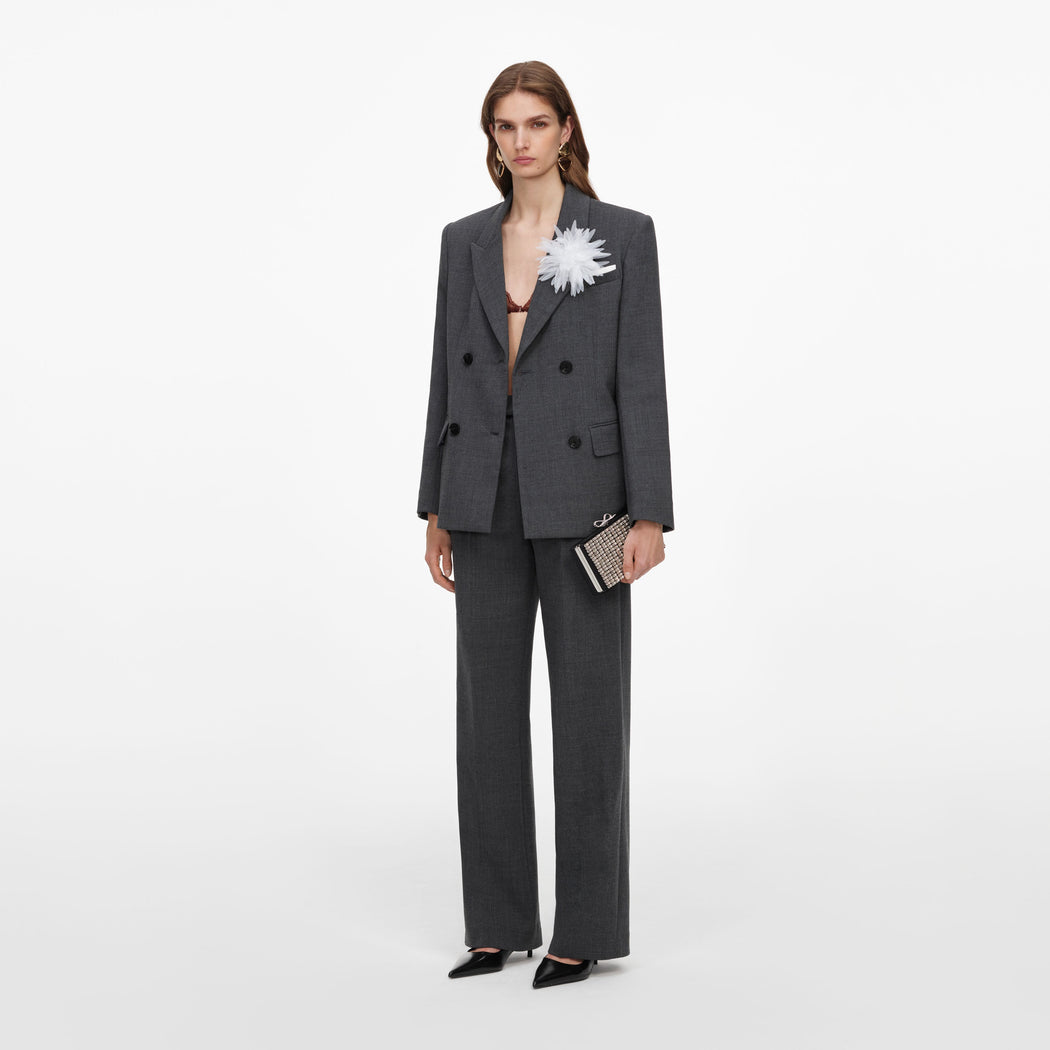 Woman in a dark grey double-breasted suit, with a white flower pinned to the lapel, holding a clutch bag