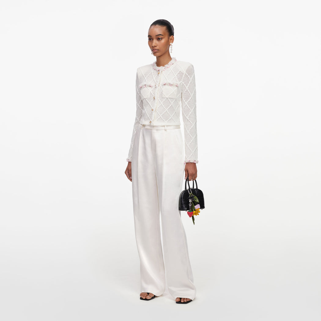 Woman in white crochet jacket with pink trim, white wide-leg trousers, and black sandals, holding a black handbag