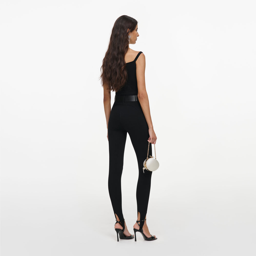 Woman in black top, leggings, and heels, holding a white circular purse