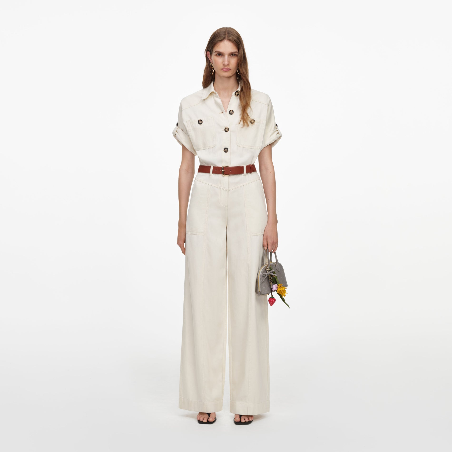 Woman in a cream short-sleeved button-up shirt, wide-leg pants, brown belt, and black sandals holding a small gray handbag