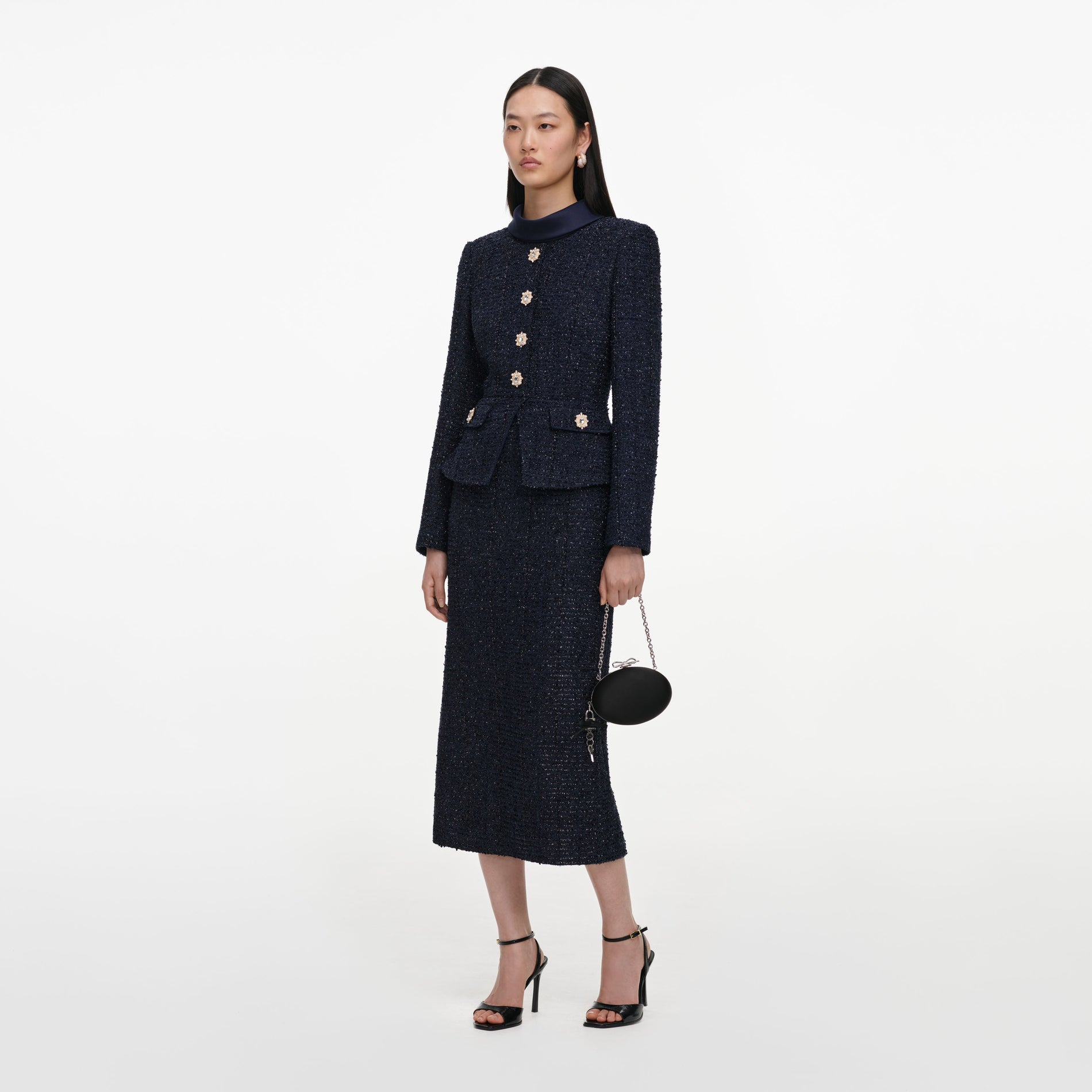 Woman in a navy blue tweed jacket with gold buttons and a matching midi skirt, holding a black clutch bag