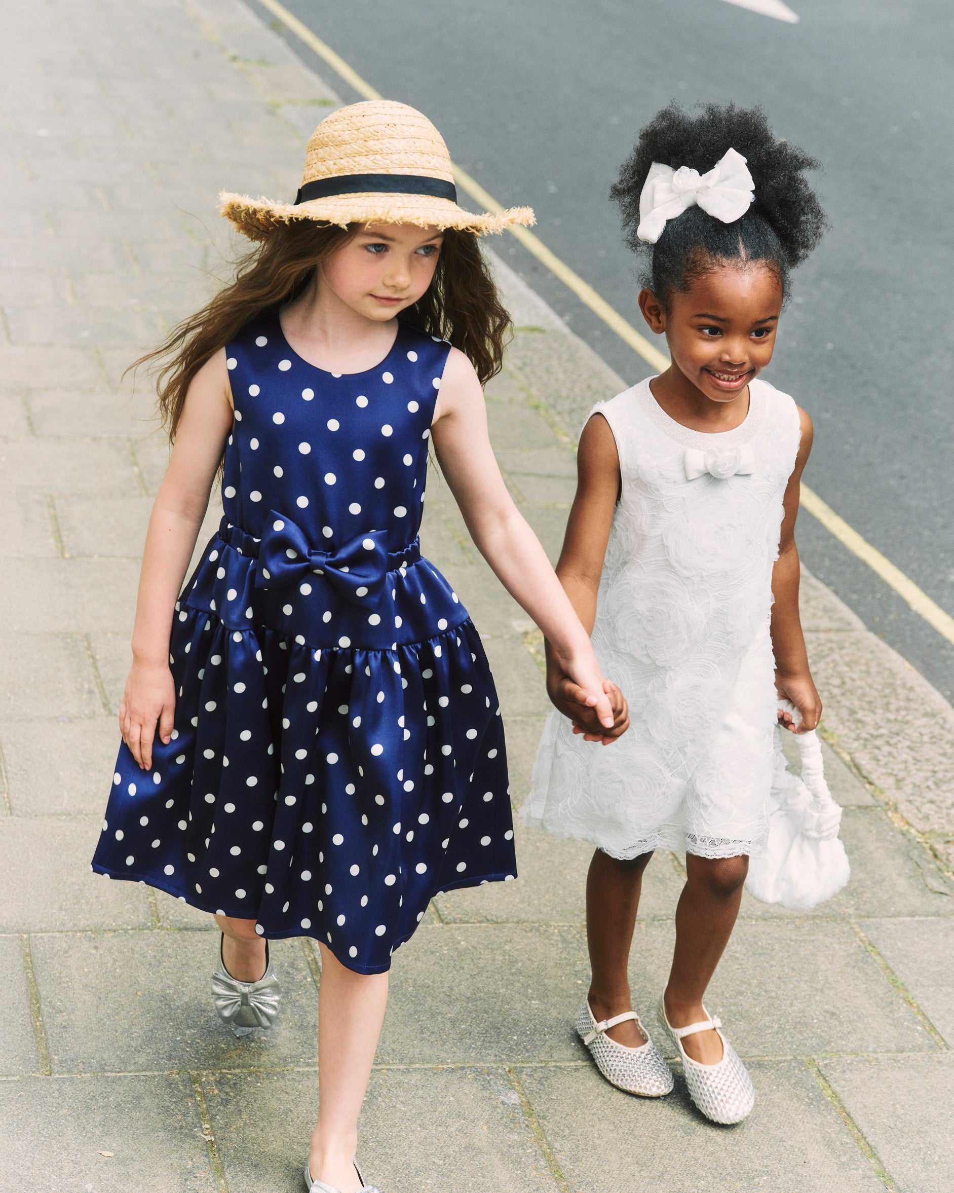 Two girls holding hands, one in a blue polka dot dress and straw hat, the other in a white dress