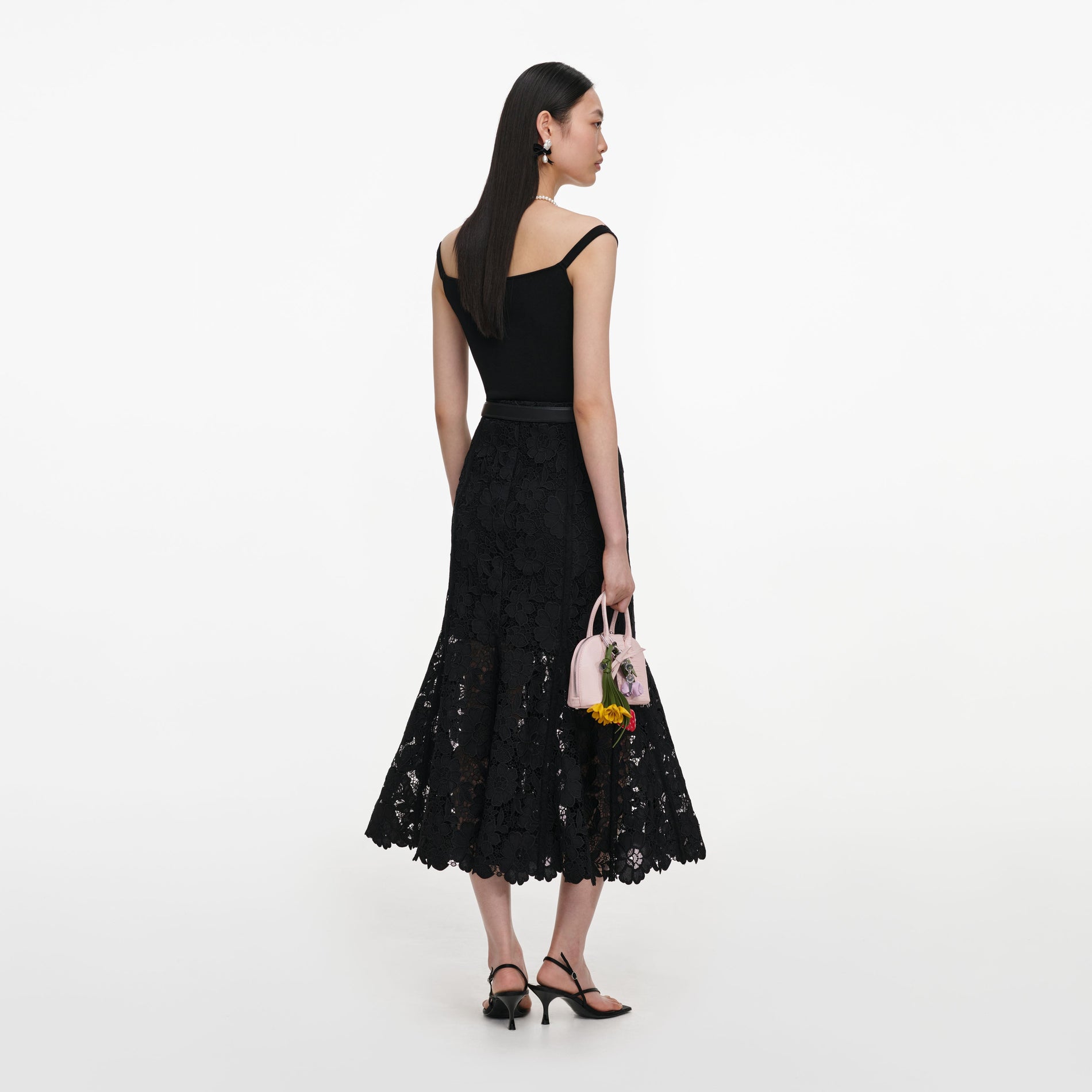Woman in black lace dress, pearl necklace, and black bow earrings, holding a pink handbag with flower charms
