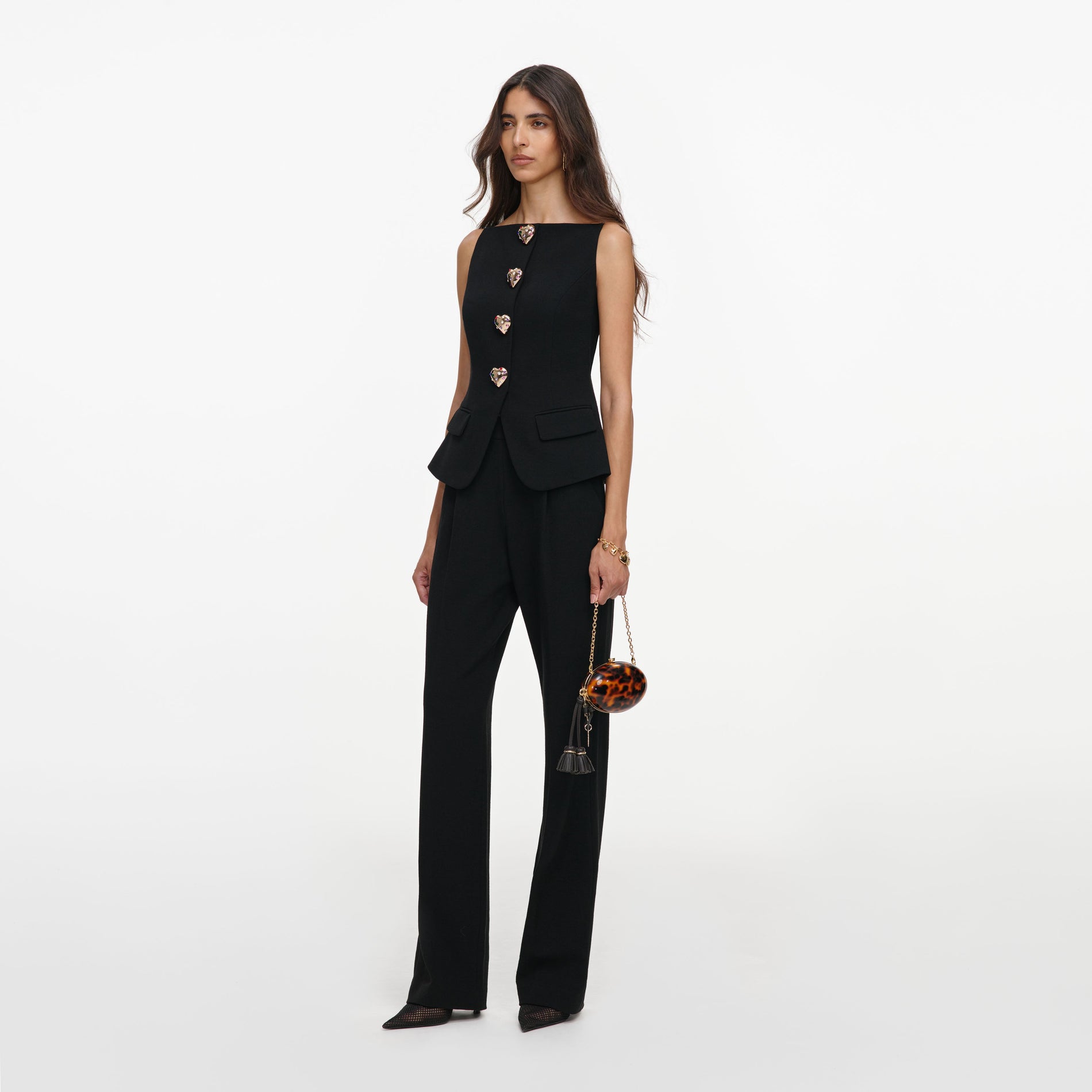 Woman in black sleeveless top with heart buttons and black trousers, holding a round tortoiseshell bag