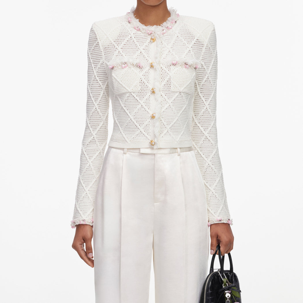Woman in white crochet cardigan with pink and pearl embellishments, white trousers, and black handbag