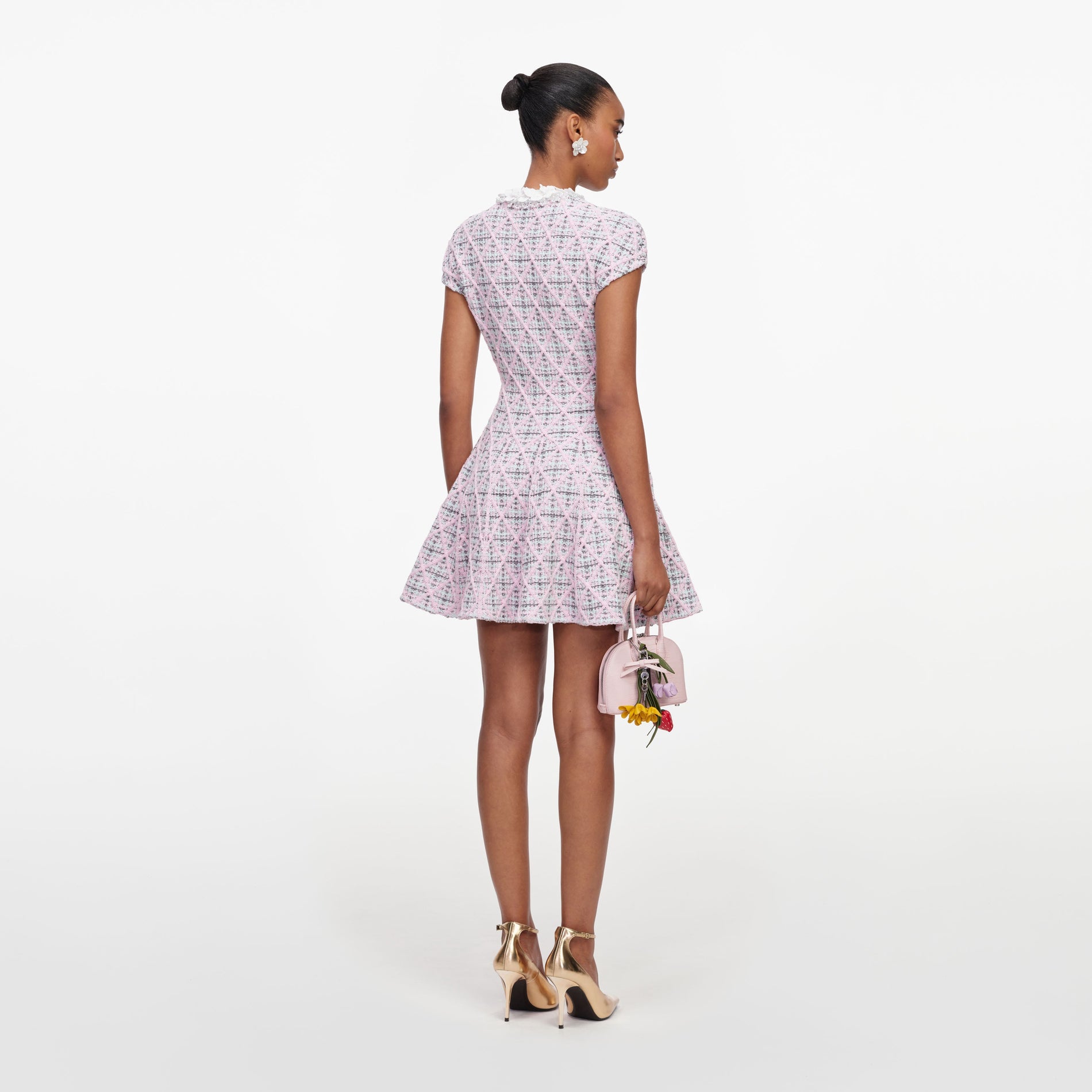 Woman in a pink and white tweed dress, gold heels, and a pink handbag with flower charms, viewed from behind