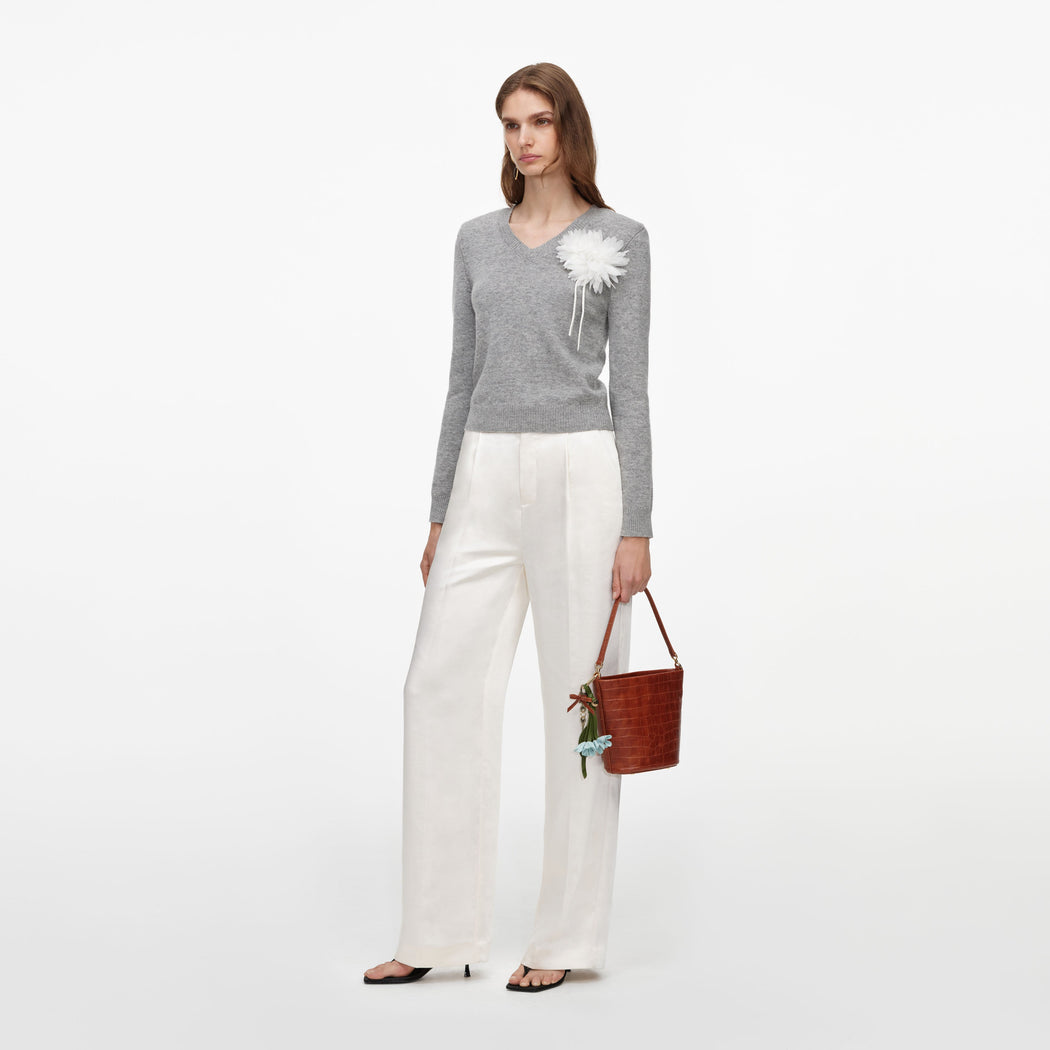 Woman in a grey v-neck sweater with a white flower brooch, white wide-leg trousers, and black heels