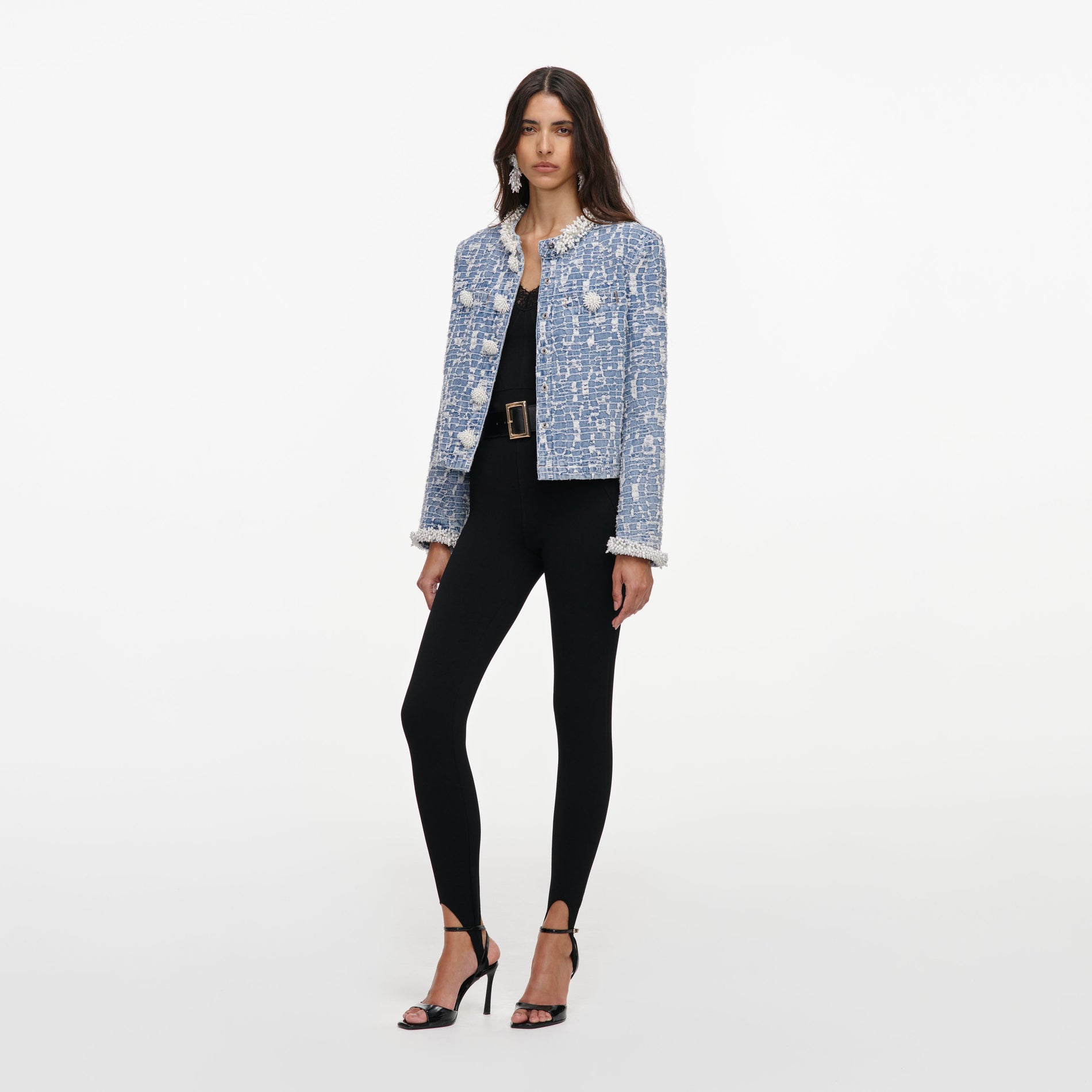 Woman wearing a blue and white tweed jacket with pearl details, black top, black leggings, and black heels
