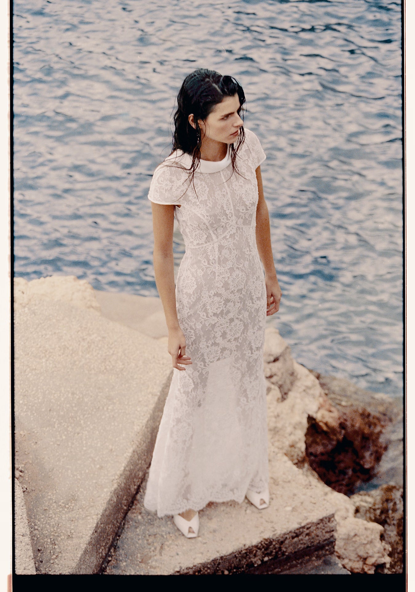 Woman in a white lace dress standing by the water.
